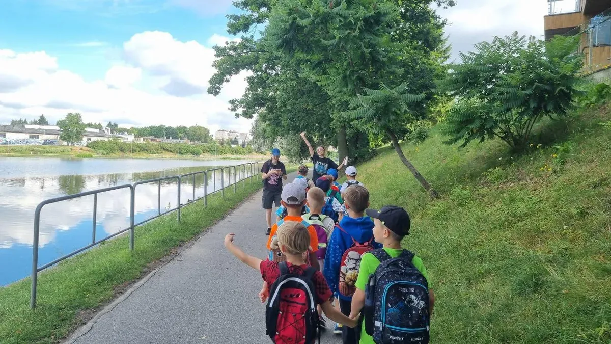 Grupa dzieci z plecakami idzie ścieżką nad wodą